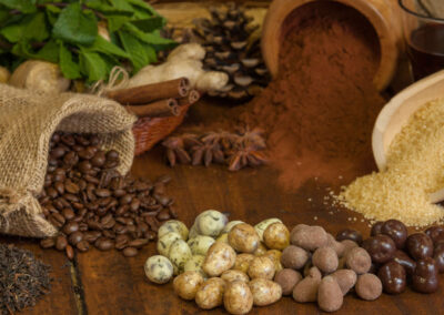 Un assortimento di ingredienti naturali e cioccolatini artigianali disposti su un tavolo rustico in legno. In primo piano ci sono cioccolatini ricoperti in vari gusti e colori: fondente, al latte, bianchi e spezzati. Dietro, sacchi e contenitori traboccano di chicchi di caffè, foglie di tè, zucchero di canna, cacao in polvere, cannella, anice stellato, zenzero fresco e foglie di menta. L’insieme trasmette un’atmosfera calda e artigianale, legata alla lavorazione del cioccolato di qualità.