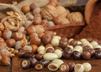 Nocciole Assortite Dragèes, nell'immagine nocciole ricoperte di cioccolato e cioccolato bianco, sullo sfondo noci e nocciole piemontesi usate nella produzione
